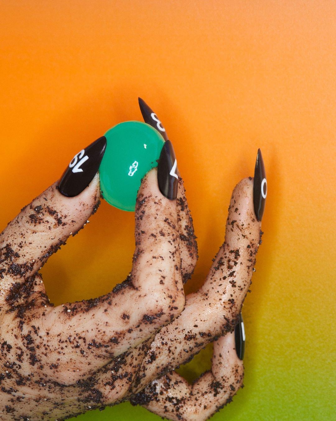 Close-up of a hand with decorated nails holding a green ball against an orange and green gradient background. Manicured nails are black with white text.