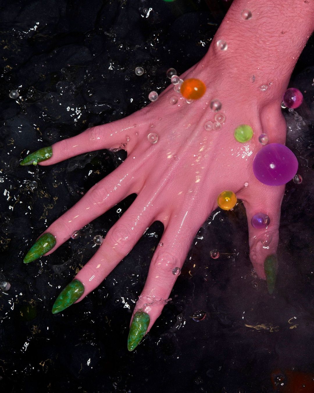 Close-up of a pink hand with green nails in dark water, adorned with colorful, translucent gel beads, showcasing beauty and unique style.