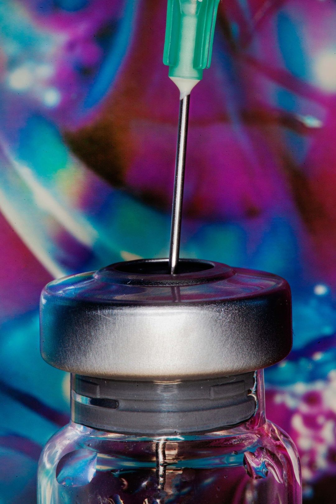 Close-up of a needle piercing the top of a vial, set against a vibrant, blurred backdrop of blues, pinks, and purples.