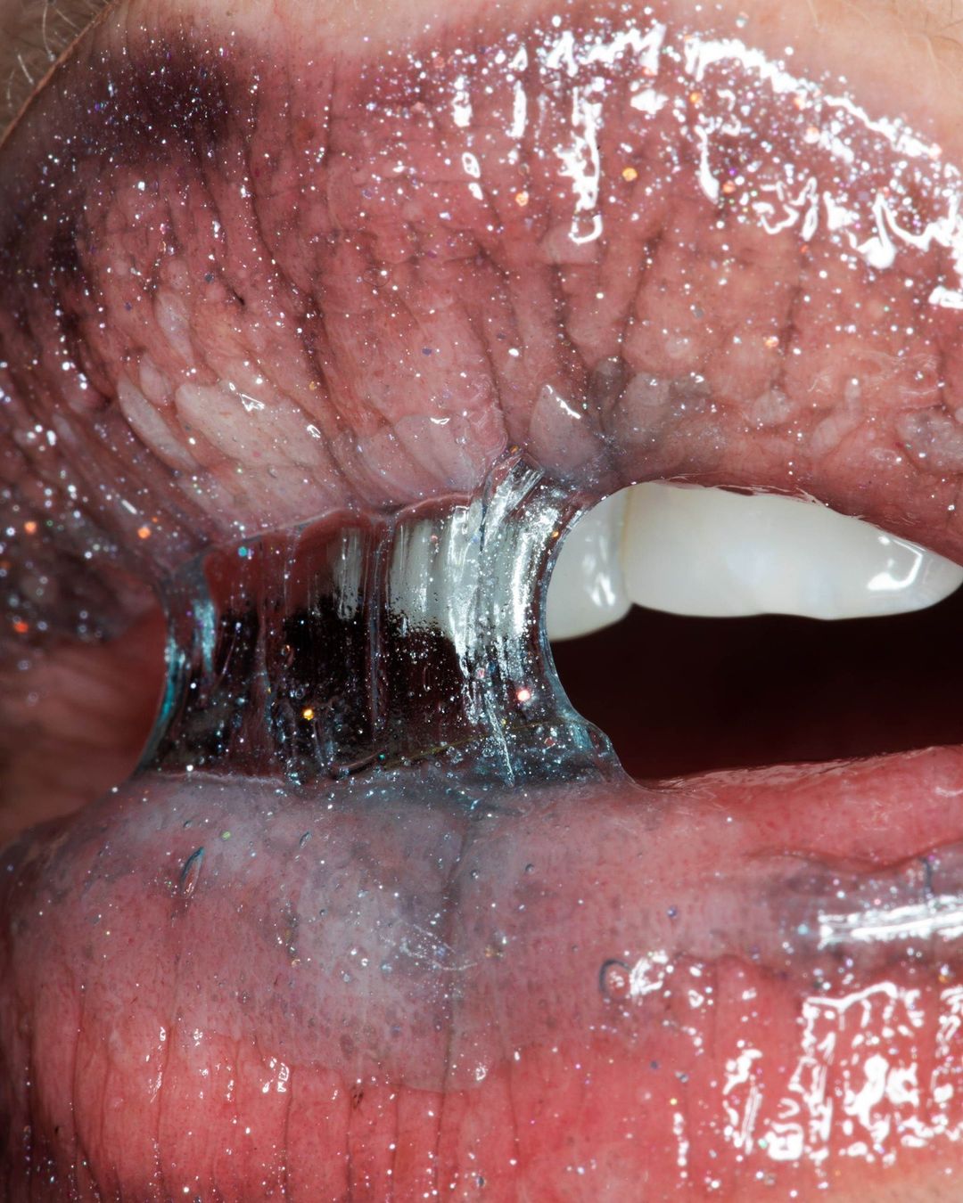 Close-up studio shot of open human mouth with teeth visible and covered with shiny blue lip gloss with glitter accents.
