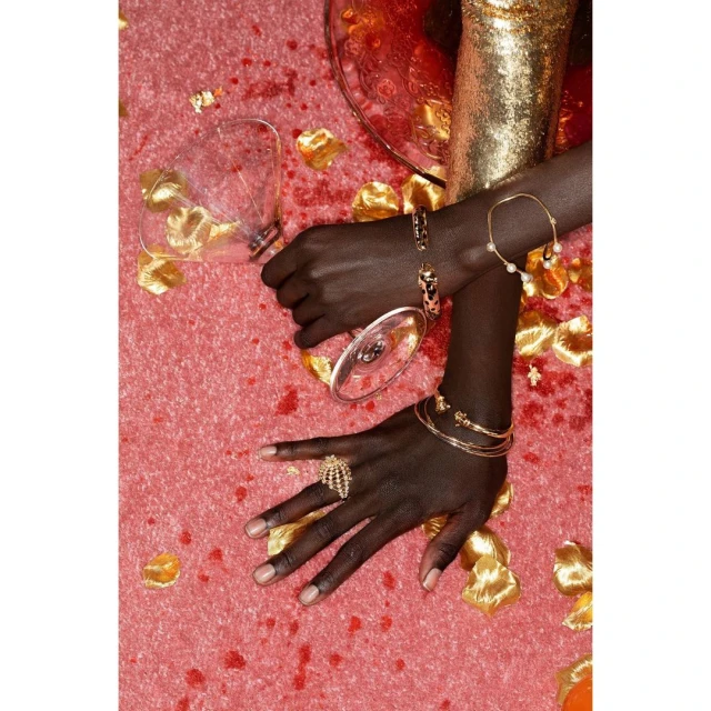 Close-up shot of a person's hands adorned with gold jewelry on a pink surface scattered with rose petals and a tipped wine glass.