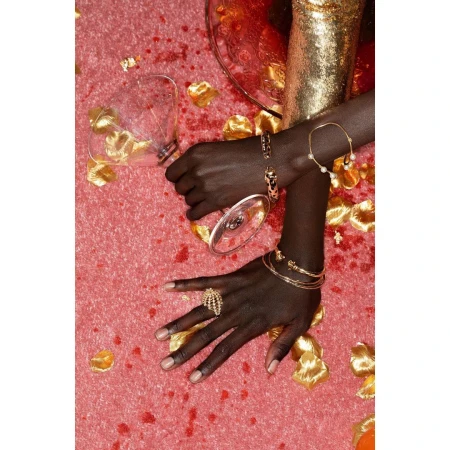 Close-up shot of a person's hands adorned with gold jewelry on a pink surface scattered with rose petals and a tipped wine glass.