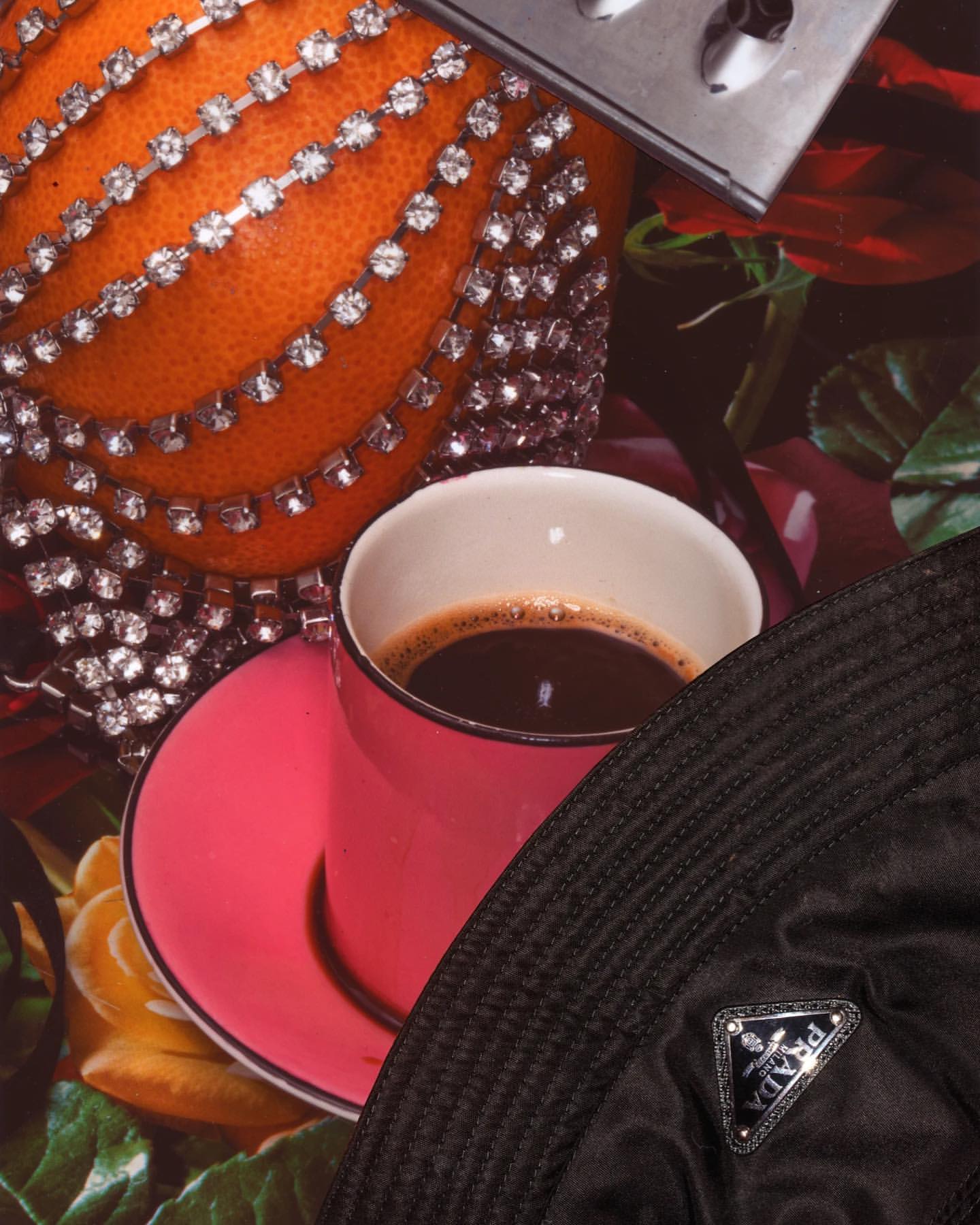 Still life with coffee in a pink cup on a saucer, bejeweled orange, metal grater, Prada bucket hat, and floral background.