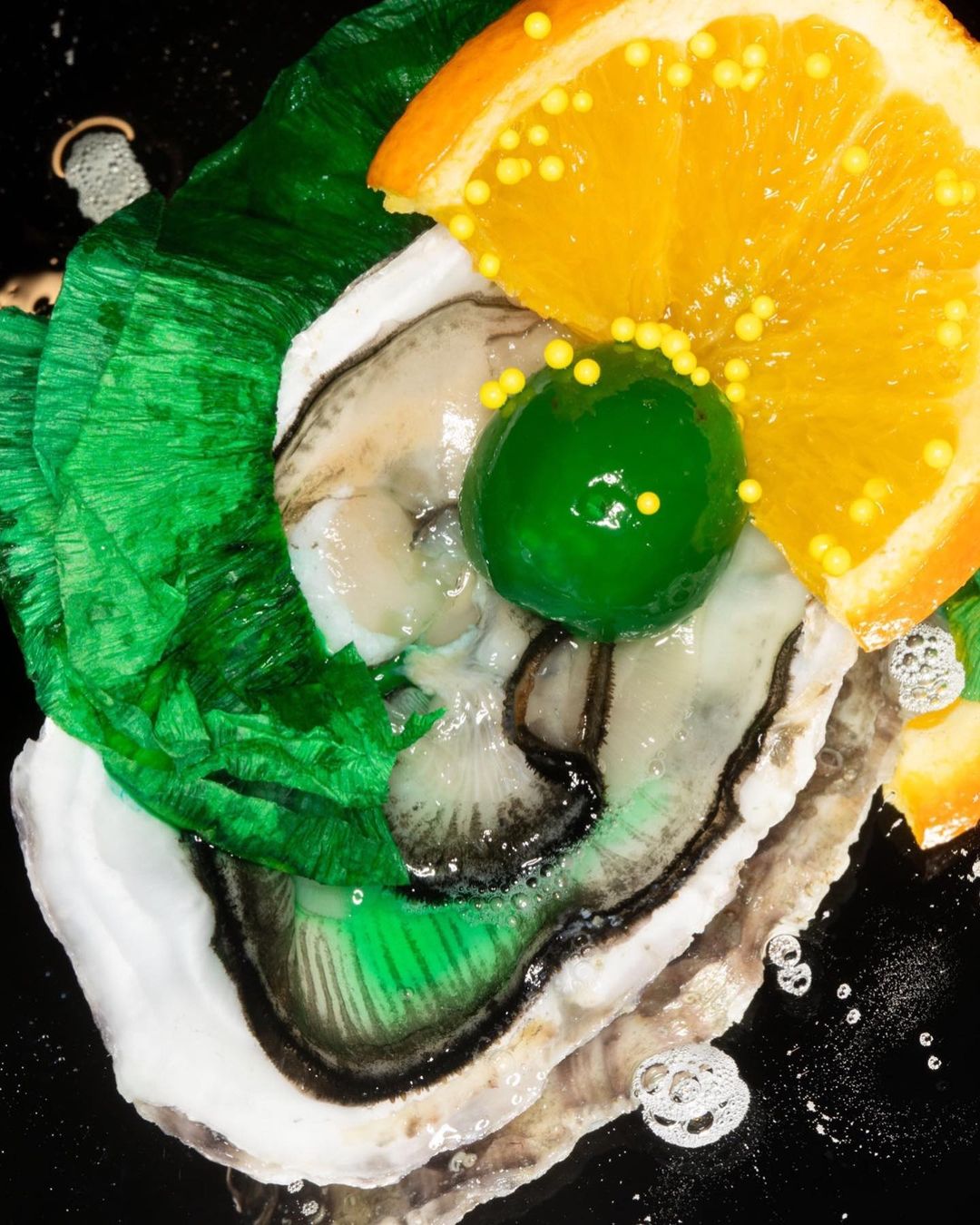 Overhead shot of an oyster topped with an orange slice, green candied fruit, and yellow sprinkles on a black surface, creating a vibrant culinary display.