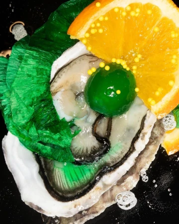 Overhead shot of an oyster topped with an orange slice, green candied fruit, and yellow sprinkles on a black surface, creating a vibrant culinary display.