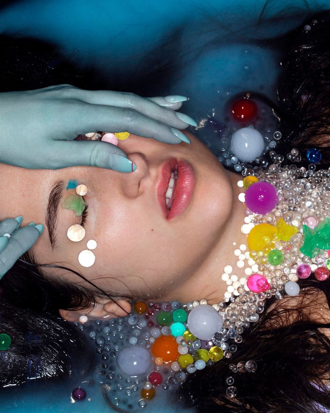Conceptual beauty shot of a fair-skinned woman partially submerged in water with blue-painted hands and face adorned with colorful beads.
