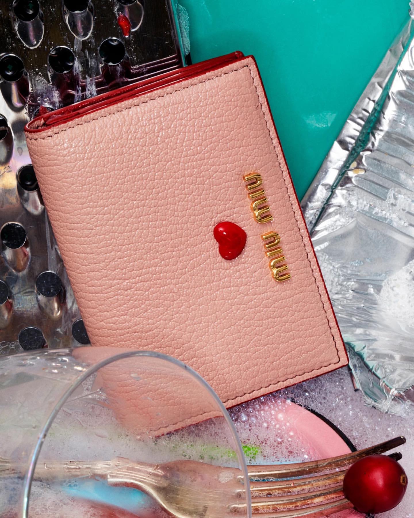 Pale pink leather wallet from Miu Miu with a red heart detail, surrounded by silverware and dishwashing detritus in a still life.
