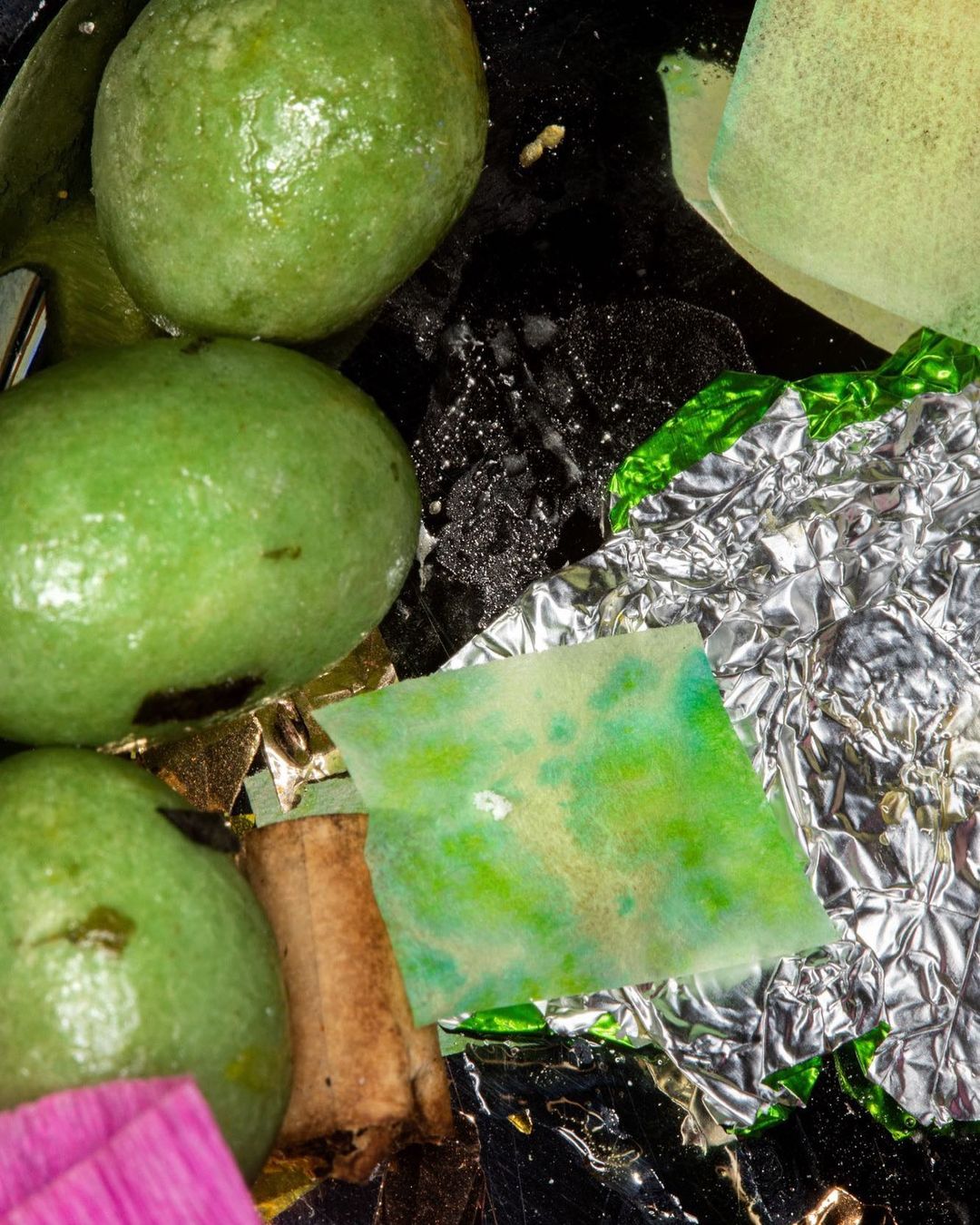 Close-up of green mochi balls, foil wrappers, and tea bag wrappers arranged on a black surface, creating an abstract and textured food still life.