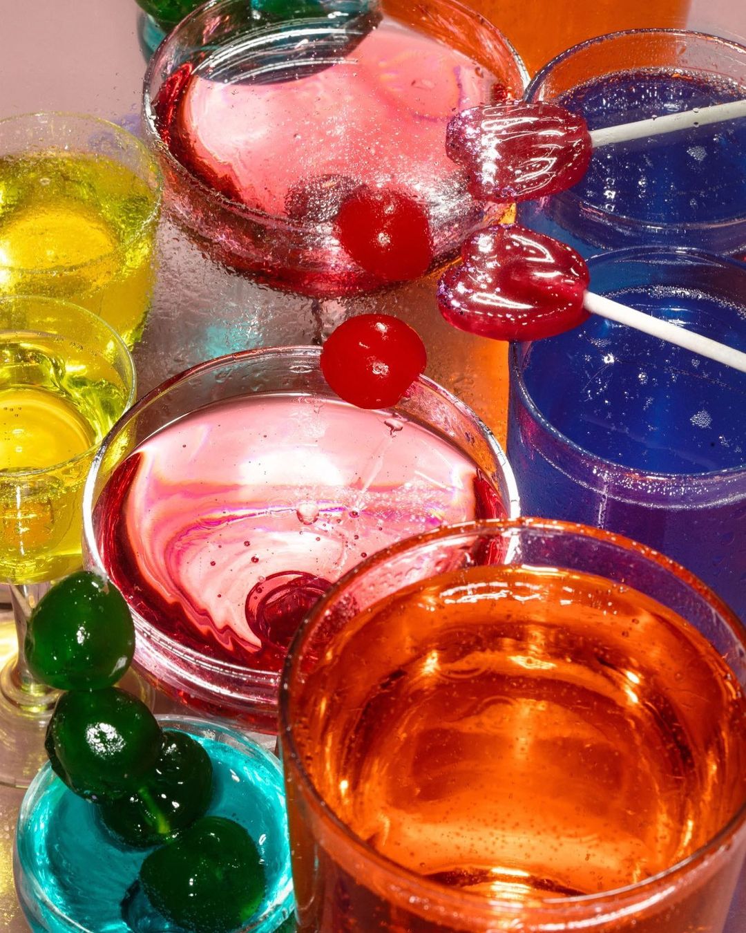 Colorful cocktail glasses with red cherries and lollipops, filled with vibrant liquids in yellow, pink, blue, and orange hues.