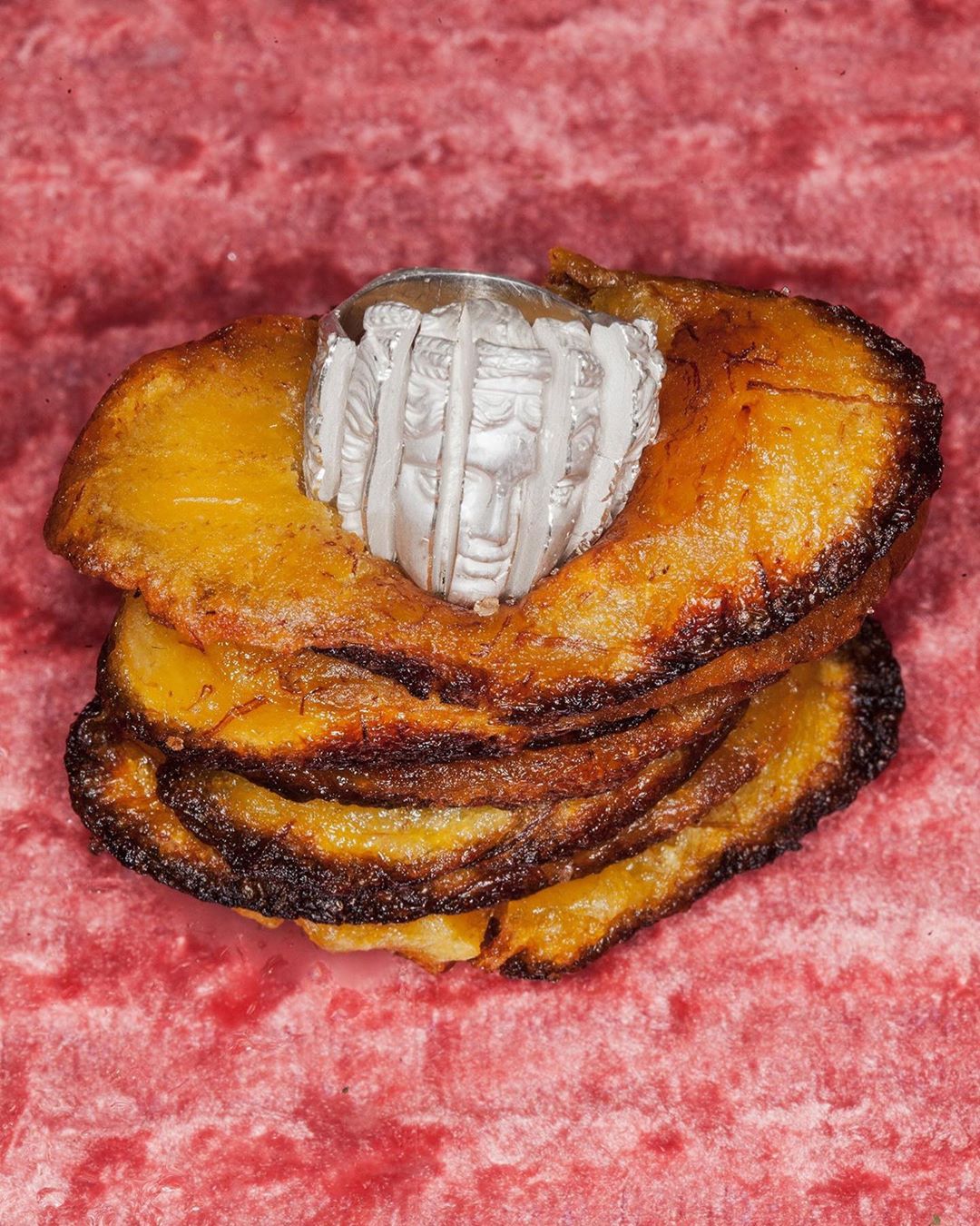 Close-up of a silver ring with face design, resting atop a stack of dried, golden-brown fruit slices against a textured pink background.