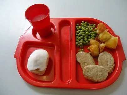 Red cafeteria tray features cubed potatoes, green peas, processed meat, iced cake, and beverage, suggesting a school lunch. Overhead view.