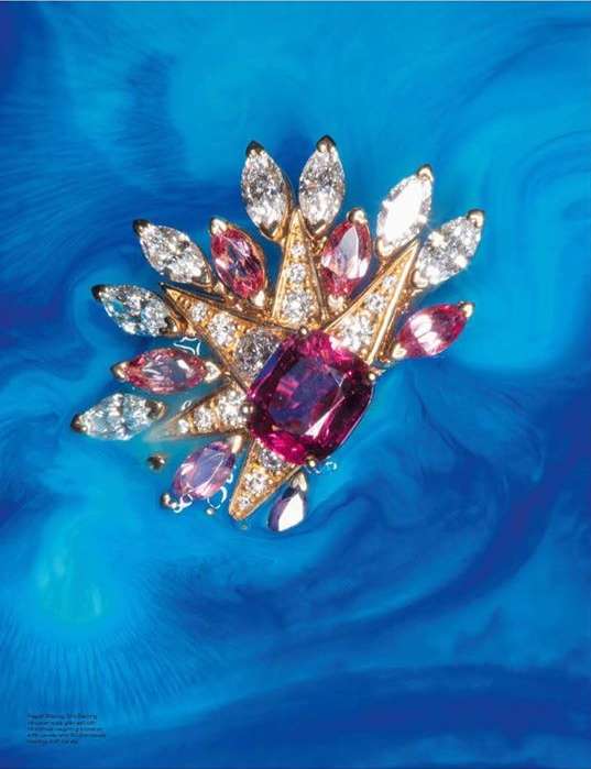 Close-up studio shot of a gold-tone floral brooch with white and pink gemstones set against a swirling teal blue background.