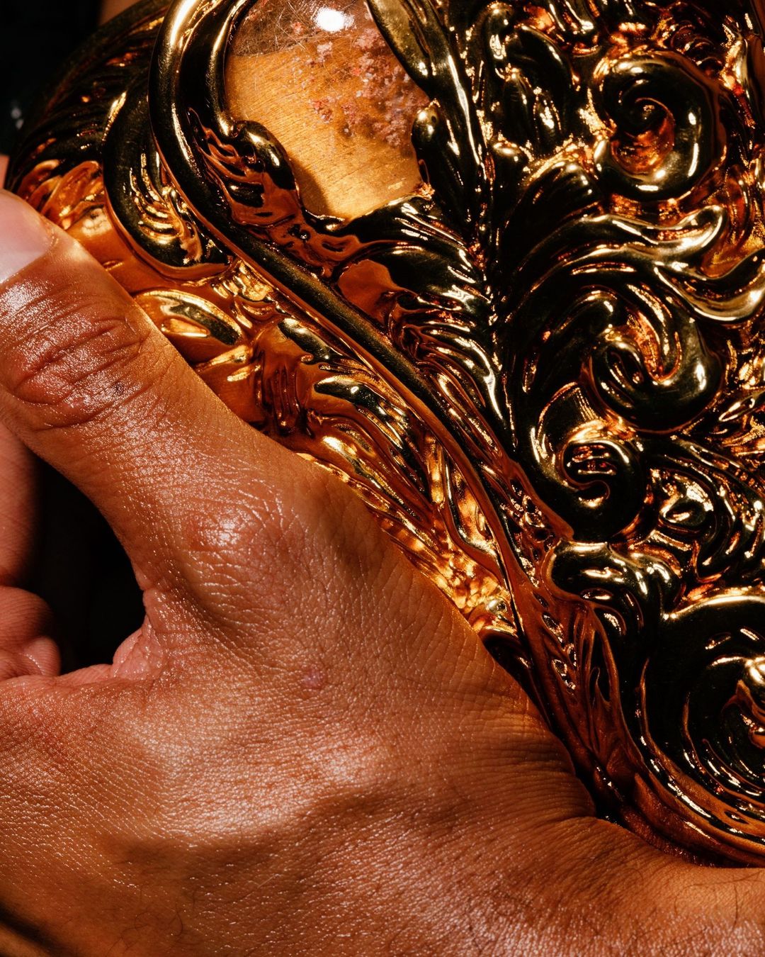 Close-up on a hand holding an ornately decorated gold object, showcasing its intricate details and reflective surface, with warm lighting.