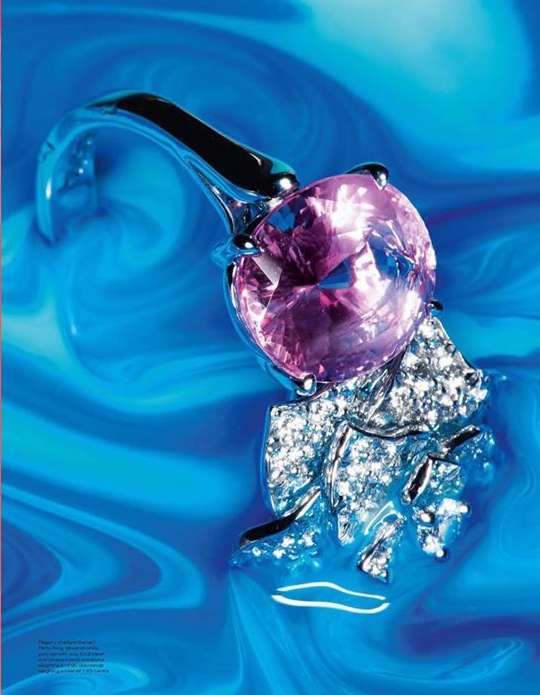 Close-up studio shot of a jewelry ring with a large round pink gem, silver band, and diamond setting against a swirling blue background.