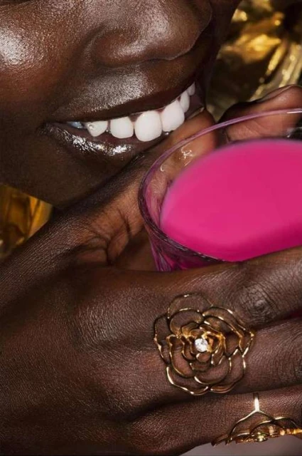 Close-up of a dark-skinned person smiling, wearing flower-shaped rings, and holding a pink drink in a clear glass in a studio setting.