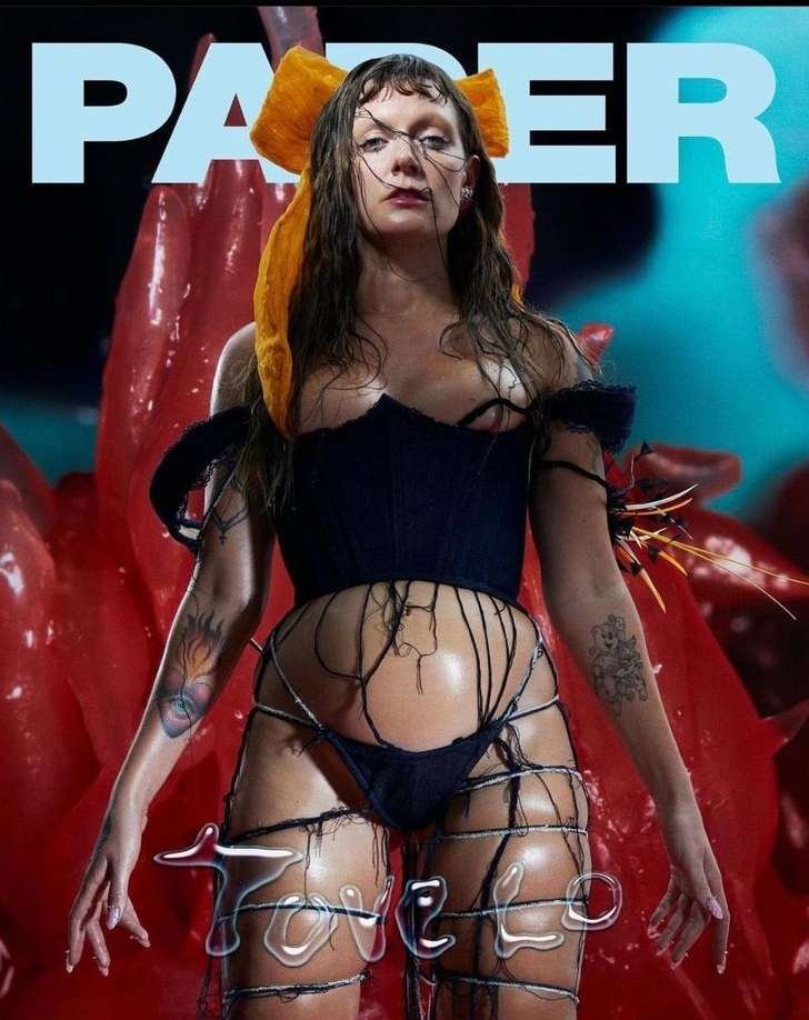 Singer Tove Lo poses in a low-angle shot for PAPER magazine cover, showcasing a black corset and intricate legwear against a red backdrop.