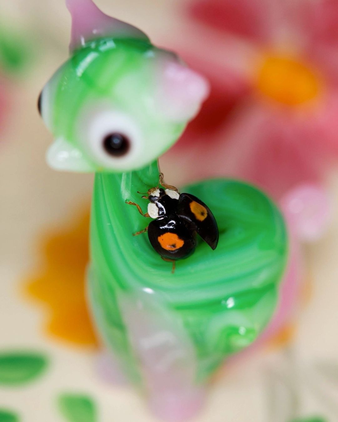 Close-up of a black-and-orange ladybug perched on a whimsical green-and-pink glass sculpture against a floral background, macro lens focus.