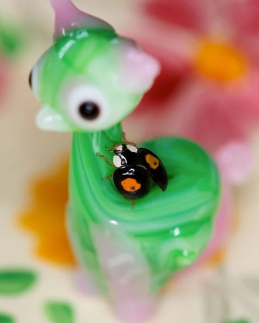 Close-up of a black-and-orange ladybug perched on a whimsical green-and-pink glass sculpture against a floral background, macro lens focus.