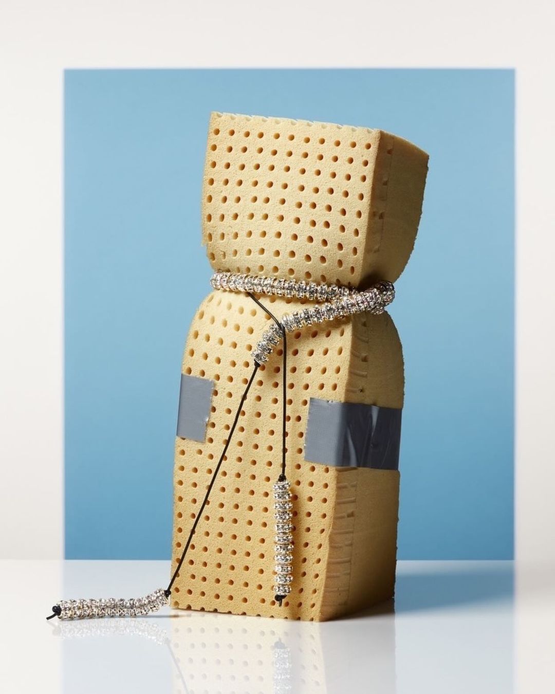 Abstract presentation of a textured block wrapped in a crystal necklace and secured with duct tape, set against a blue gradient background.