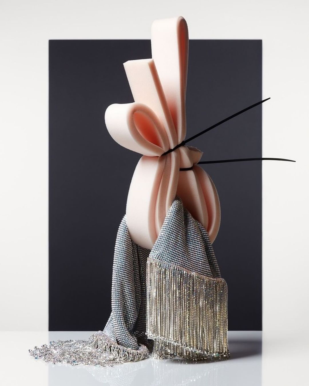 Abstract sculpture with pink foam and a fringed, silver scarf against a dark grey and white backdrop in a studio setting.