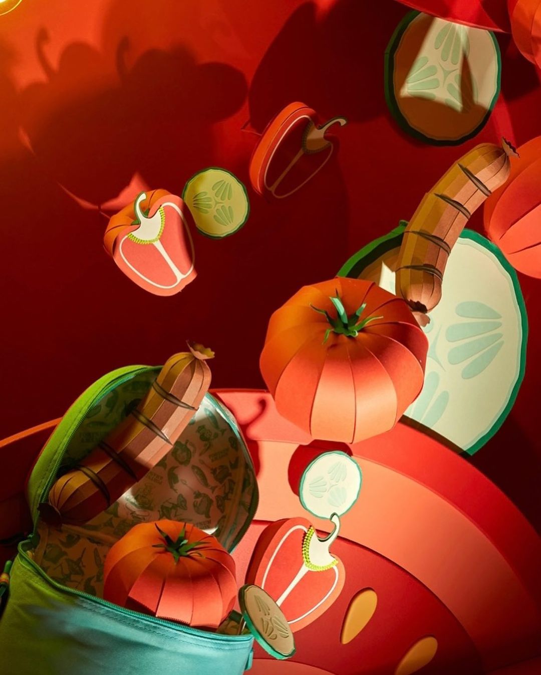 Whimsical still life of papercraft vegetables spilling from a teal bag against a bright red background with vibrant food illustration.