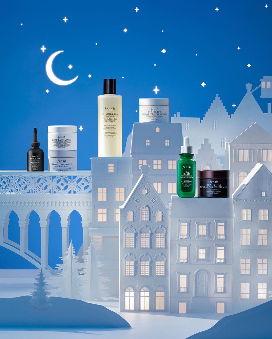 Still life of various Fresh skincare products against a backdrop of white paper cut-out buildings and a blue night sky with a crescent moon.