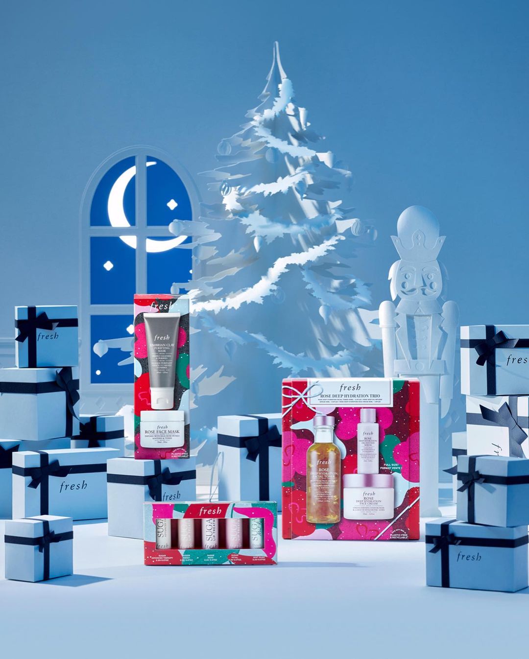 Fresh branded beauty gift sets arranged with a paper Christmas tree, paper moon window, and paper nutcracker in a cool-toned product shot.