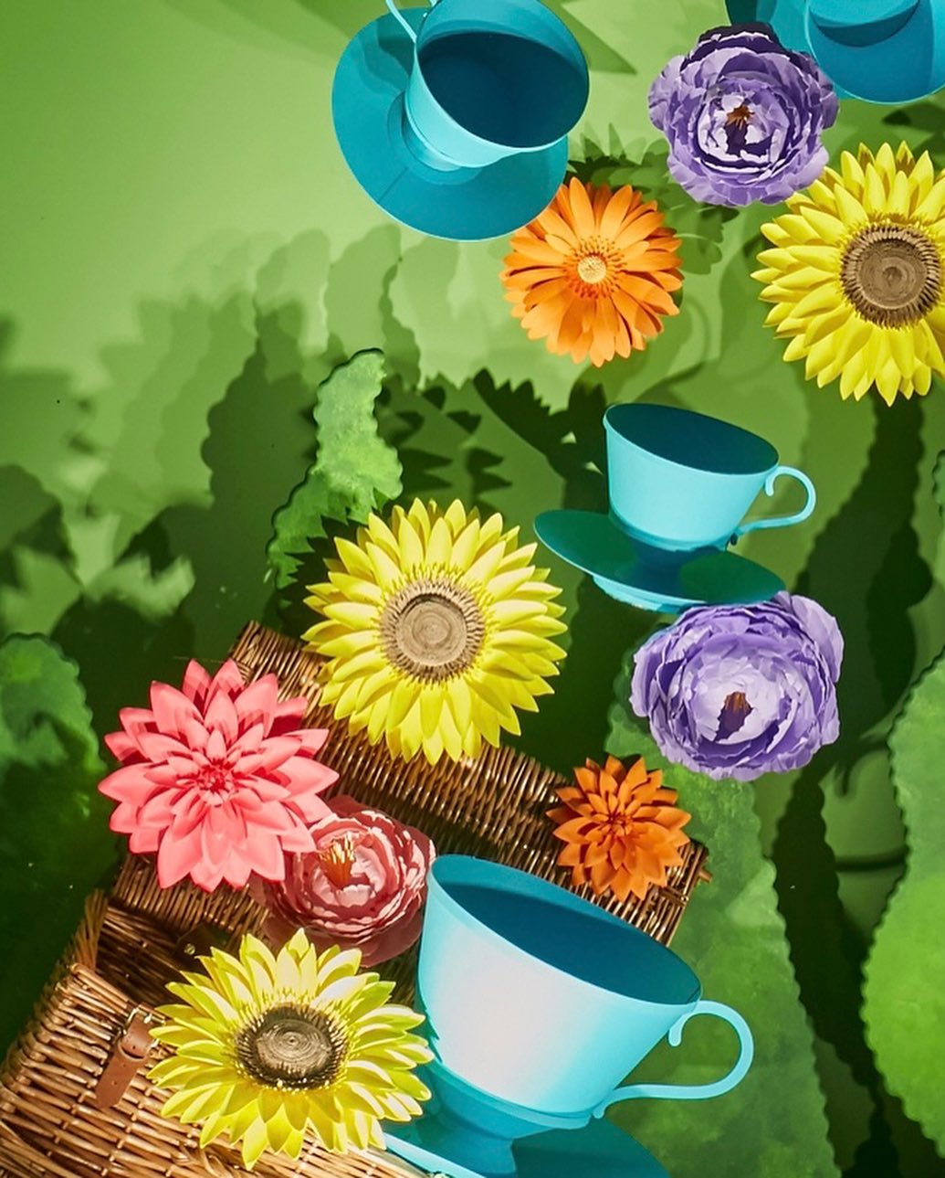 Artistic still life of tea cups and colorful flower blossoms arranged on a green background with a woven picnic basket detail.