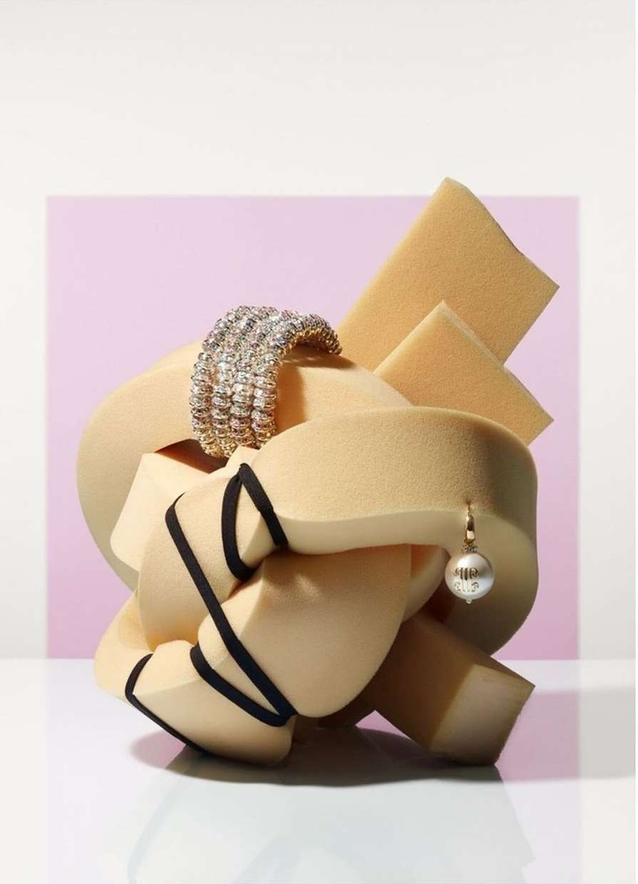 Studio shot of a silver bracelet, ball earring, and black cords tangled in peach-colored foam against a light pink and white background.