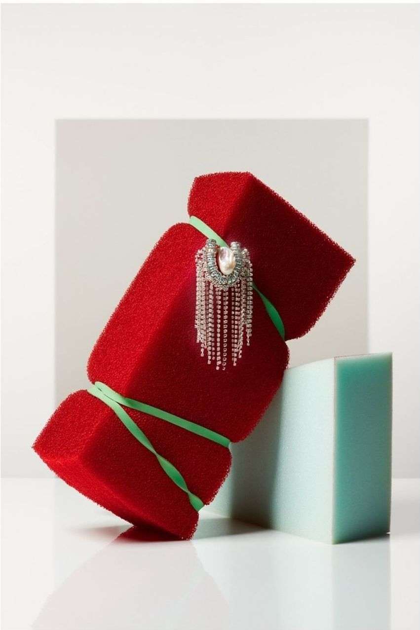 Still life of two red sponge blocks connected by green rubber bands, with an ornate silver and pearl brooch on a white reflective surface.