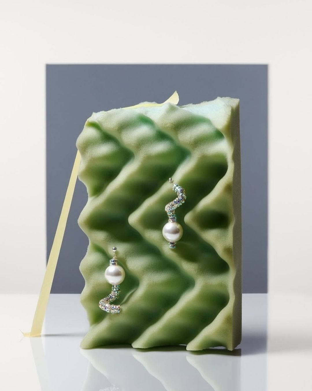 Elegant pearl earrings displayed on textured green block with soft yellow ribbon against grey and white backdrop, showcasing jewelry design.