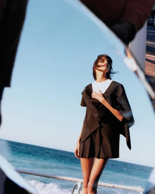 Fashion shot of an adult female model posing in a dark outfit on a sunny day by the sea, reflected in shards of broken mirror.