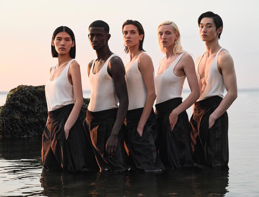 Five diverse models stand in water wearing white tank tops and dark trousers, creating a minimalist fashion editorial with a natural, serene atmosphere.