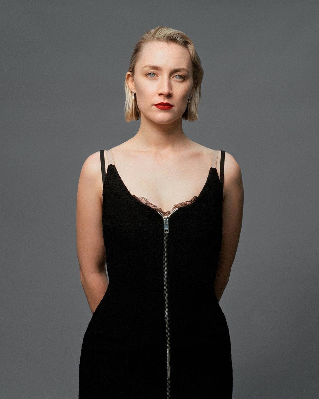 Portrait of fair-skinned woman with blonde bob wearing a black dress with a zipper, posing in a studio with a gray background.