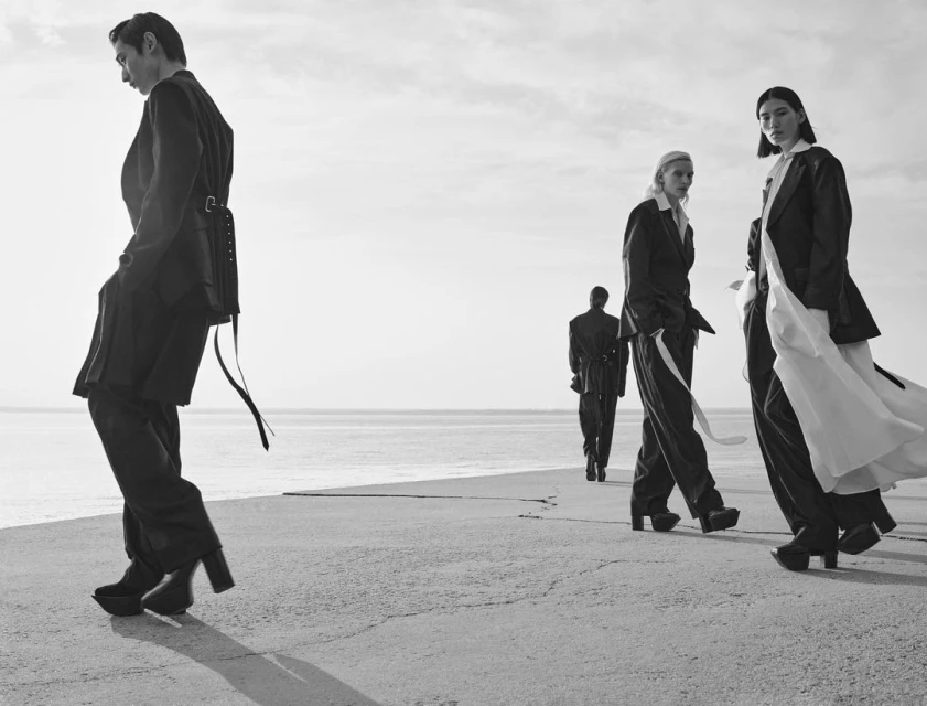 A fashion shot with five people walking on a concrete surface near a sea or large lake with a cloudy sky in the background.