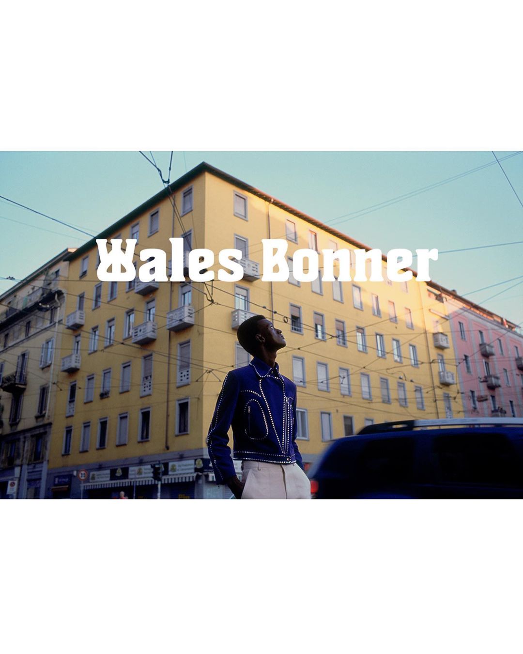 Editorial fashion shot of a Black male model in a blue jacket and cream trousers outdoors in an urban setting with buildings and a vehicle.