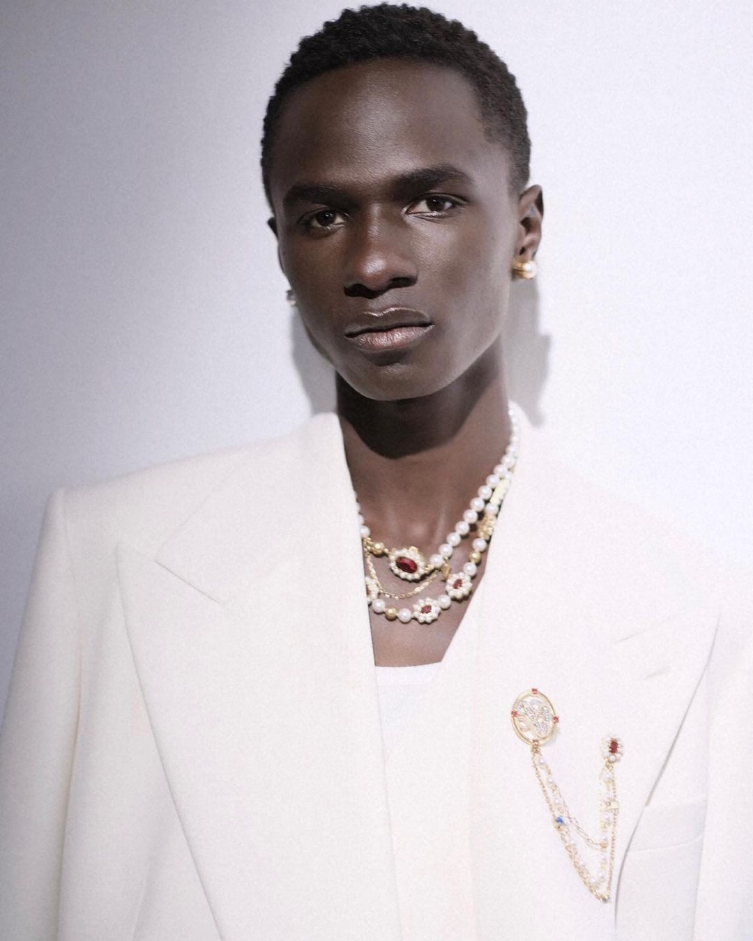 Elegant portrait of a Black male model wearing a white suit and layered pearl necklaces, posing against a light gray backdrop.