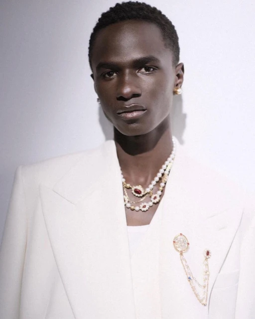 Elegant portrait of a Black male model wearing a white suit and layered pearl necklaces, posing against a light gray backdrop.