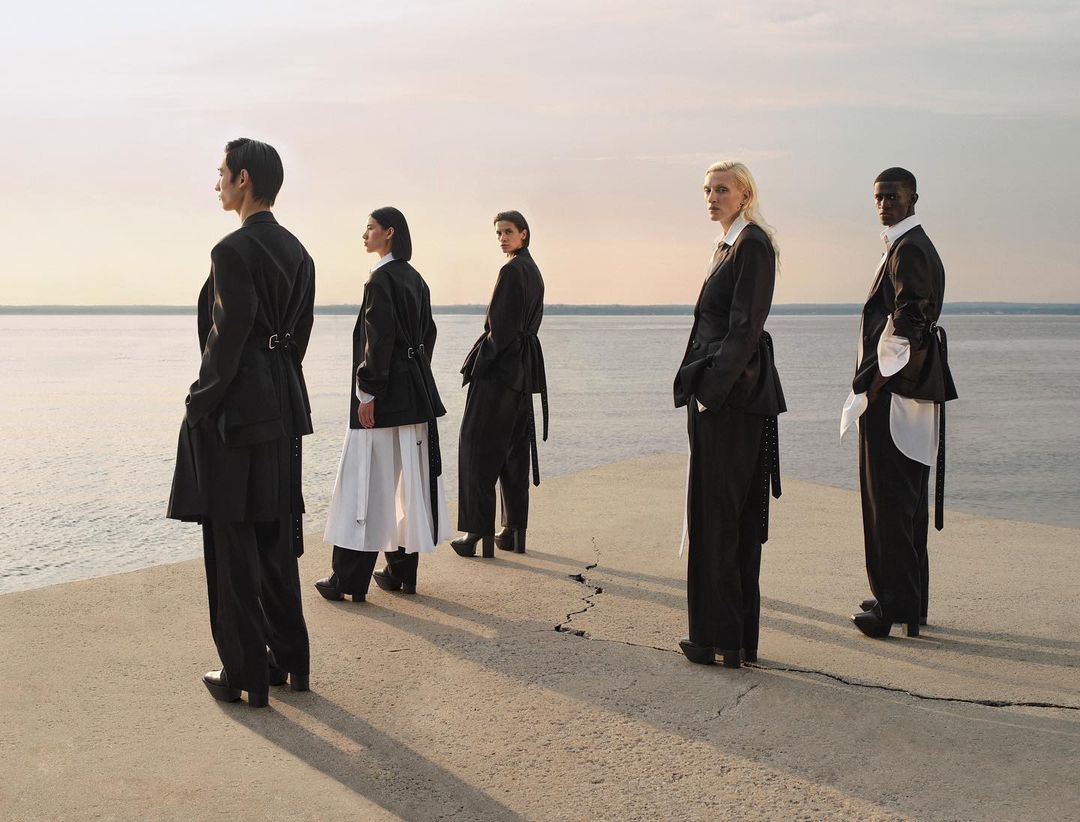 Five fashion models stand in a line outdoors overlooking a body of water, wearing black and white clothing in a scenic daytime fashion editorial.