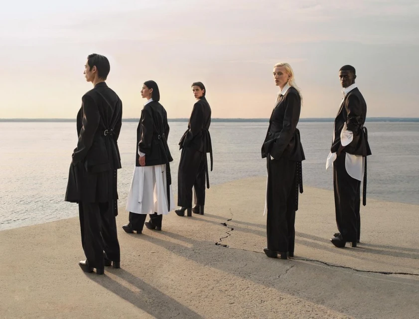Five fashion models stand in a line outdoors overlooking a body of water, wearing black and white clothing in a scenic daytime fashion editorial.