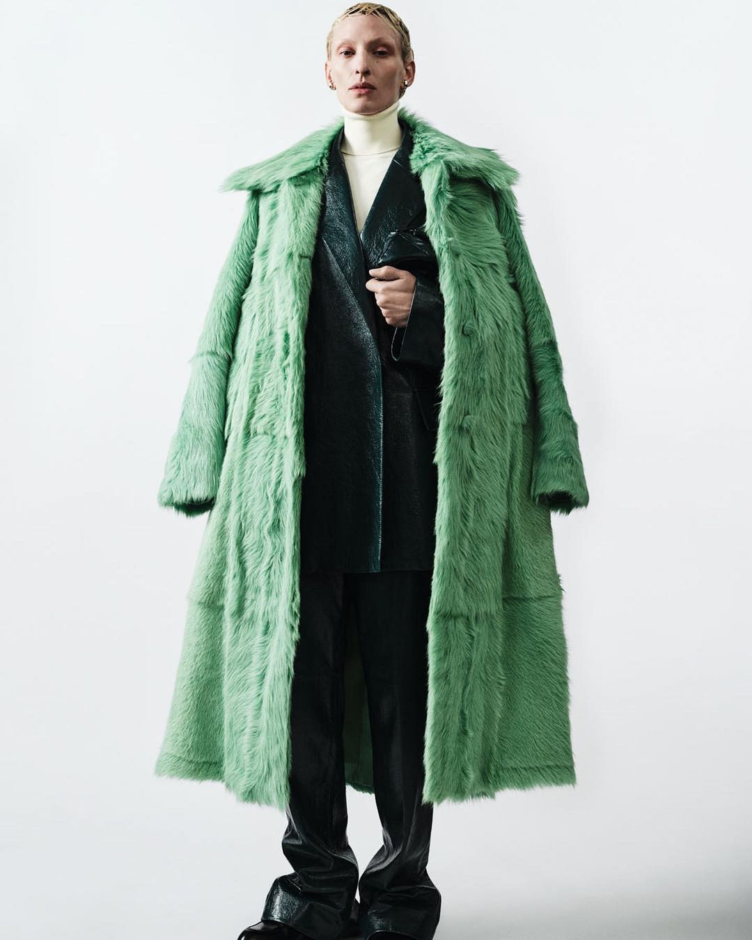 Androgynous fashion model in long green fur coat and black leather suit posing in a photo studio against a white background.