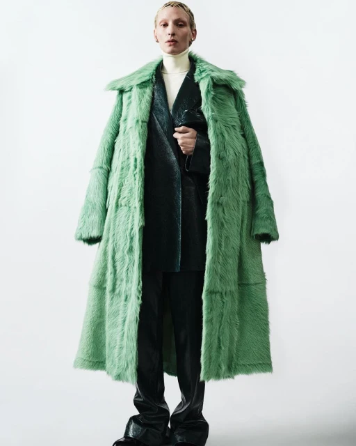 Androgynous fashion model in long green fur coat and black leather suit posing in a photo studio against a white background.