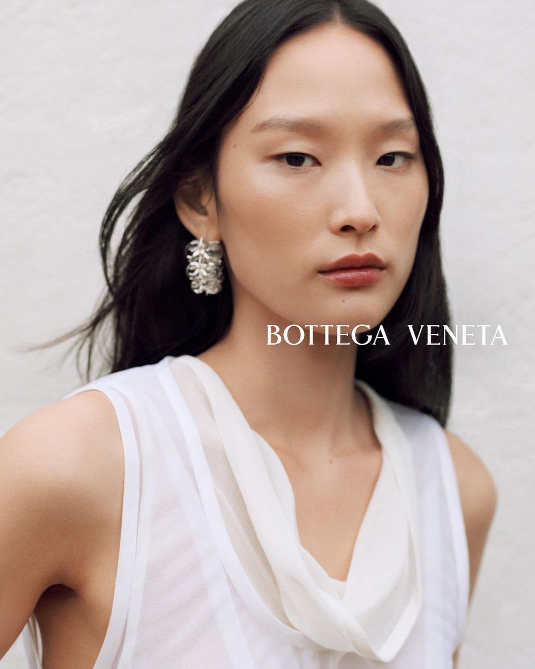Close-up portrait of an East Asian woman wearing a white top and statement silver earrings against a textured white wall.