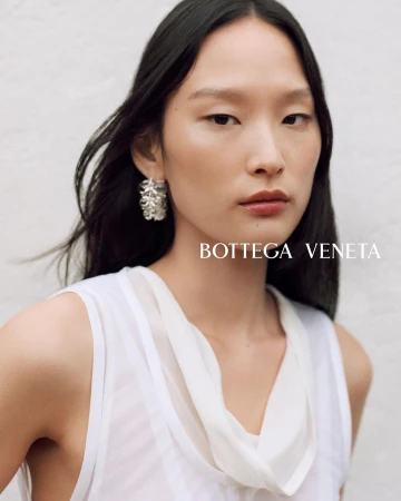 Close-up portrait of an East Asian woman wearing a white top and statement silver earrings against a textured white wall.