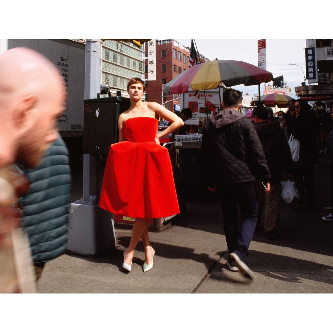 A woman in a red dress poses outdoors in an urban setting, surrounded by blurred pedestrians and street structures under daylight conditions.