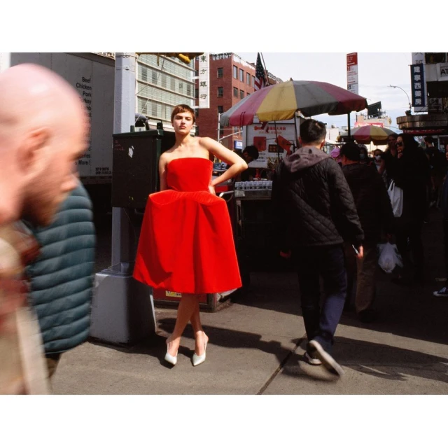 A woman in a red dress poses outdoors in an urban setting, surrounded by blurred pedestrians and street structures under daylight conditions.