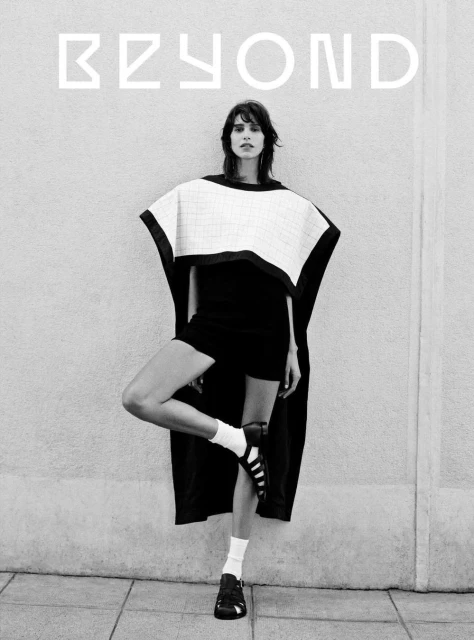 Monochrome fashion shot of a woman in dark shorts, a white top, black robe, and socks, posing against a textured wall with 'BEYOND' text.