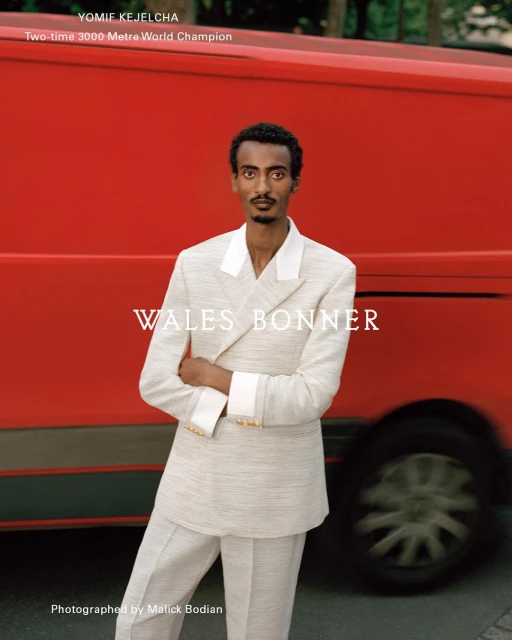 Yomif Kejelcha poses in a light-colored suit with a red van in the background, showcasing a fashion-editorial style in an outdoor setting.