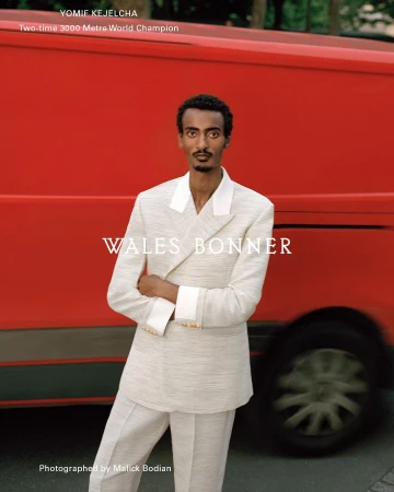 Yomif Kejelcha poses in a light-colored suit with a red van in the background, showcasing a fashion-editorial style in an outdoor setting.