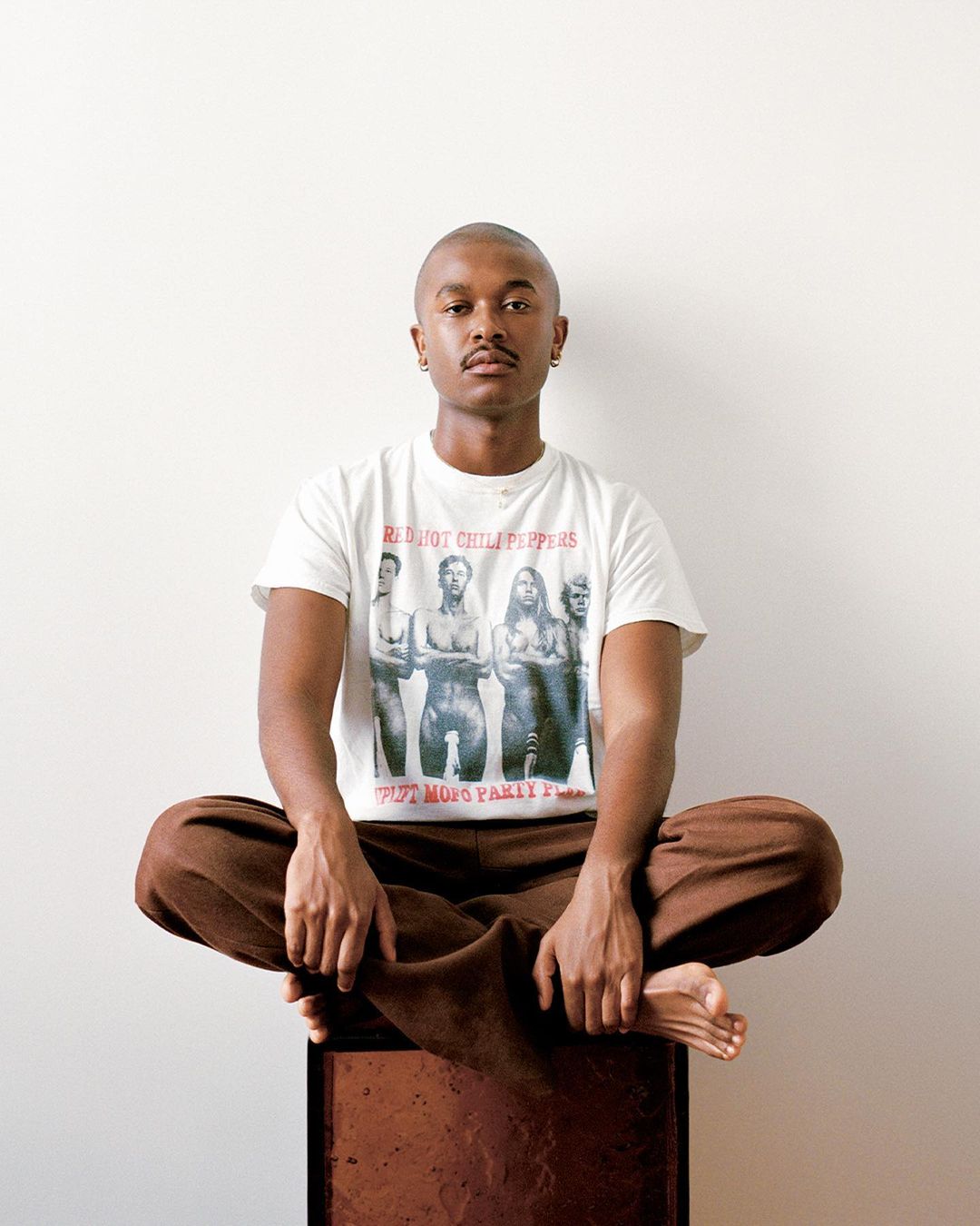 Young adult sits cross-legged on a dark box wearing a vintage Red Hot Chili Peppers t-shirt against a clean white wall backdrop.